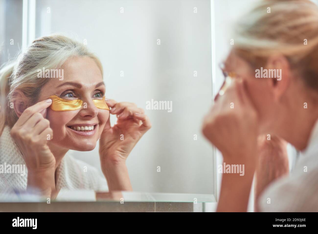 Mirror reflection of smiling middle aged caucasian woman using collagen anti aging under eye patches. Beauty, skincare and cosmetology concept Stock Photo