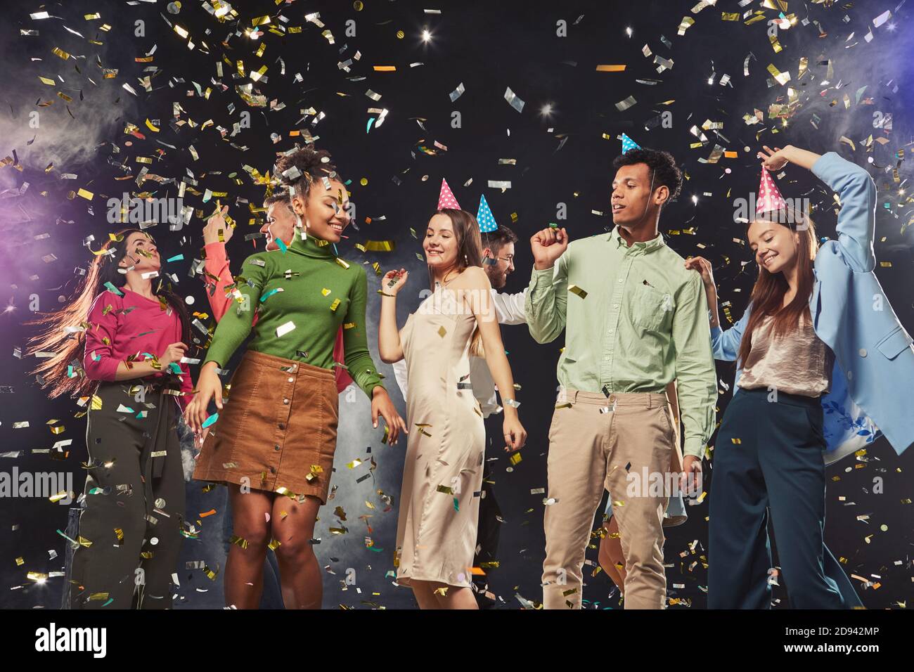 Group of young happy multiracial people wearing birthday hats and dancing, confetti falling in the air. Holiday, party and celebration concept Stock Photo