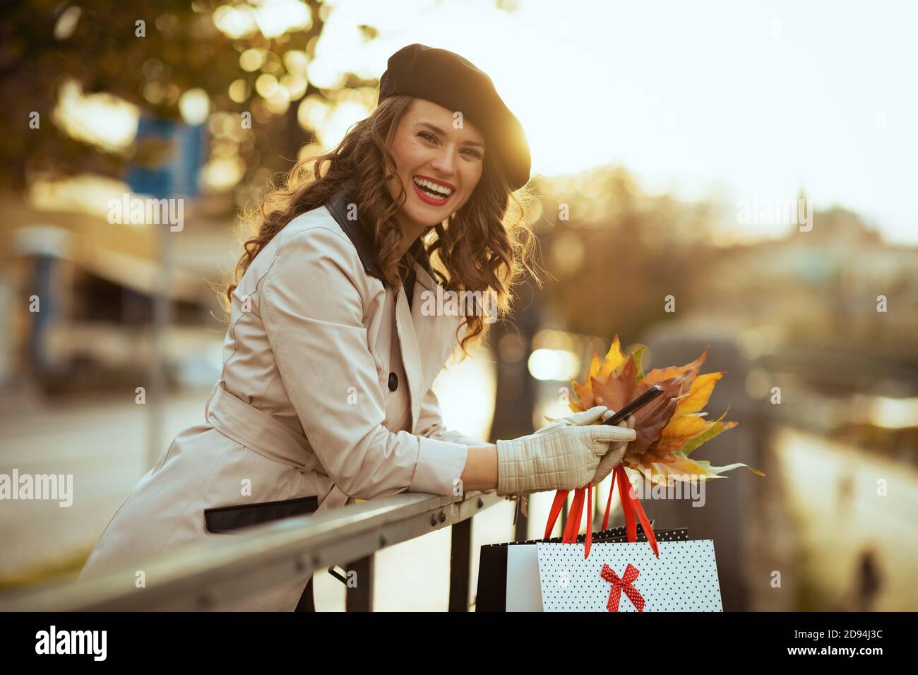 Hello autumn. smiling elegant female in beige trench coat with shopping bags and autumn yellow leaves using smartphone applications outside in the cit Stock Photo