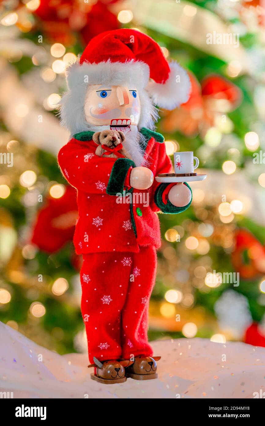 A  Santa Claus nutcracker dressed in his pajamas and teddybear and slippers holds a cup in front of a soft focused Christmas tree. Stock Photo