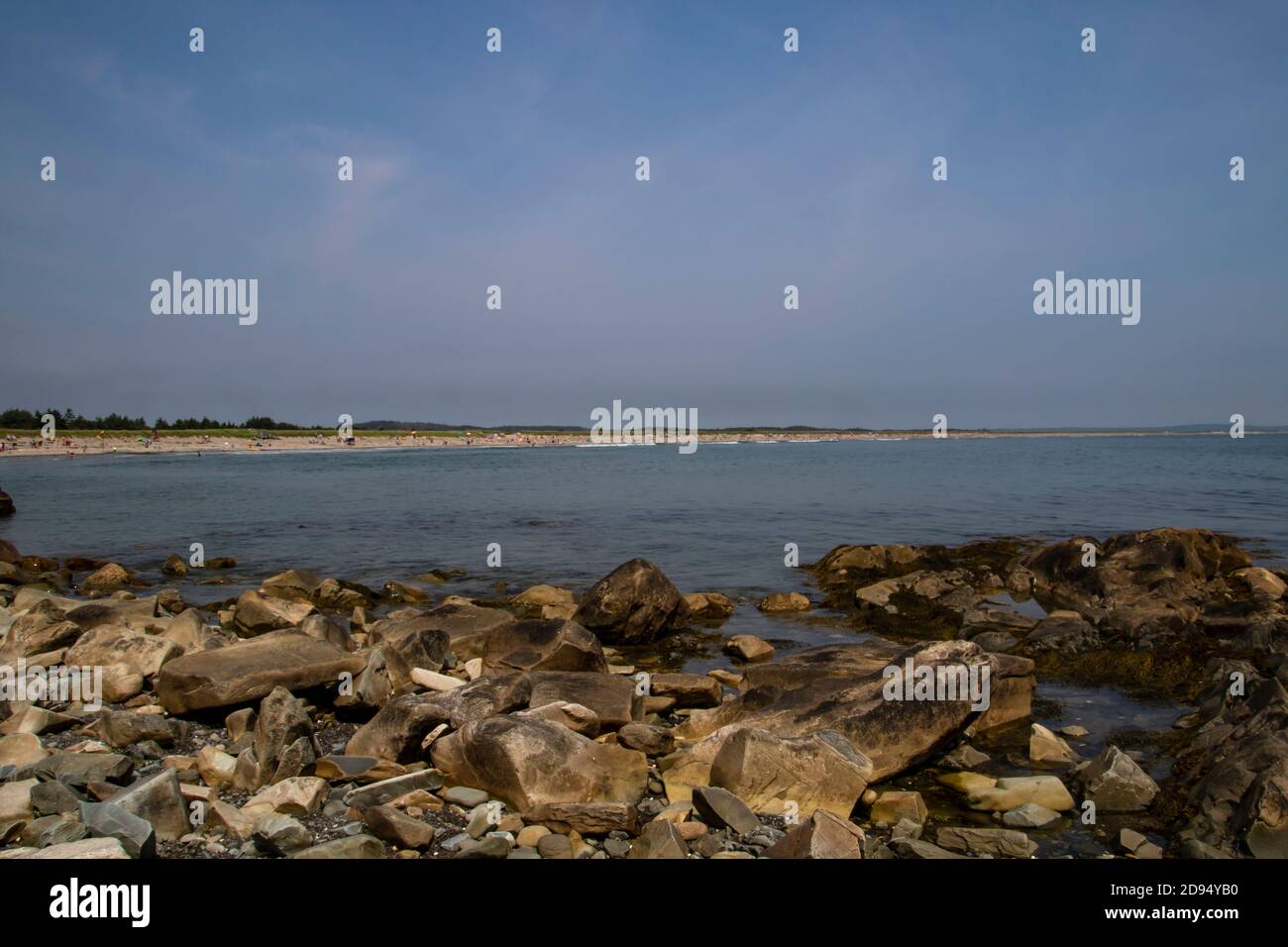 A view of a people filled beach as seen from across a bay with a rocky shoreline in the foreground.  The blue sky has an number of wispy clouds in it. Stock Photo