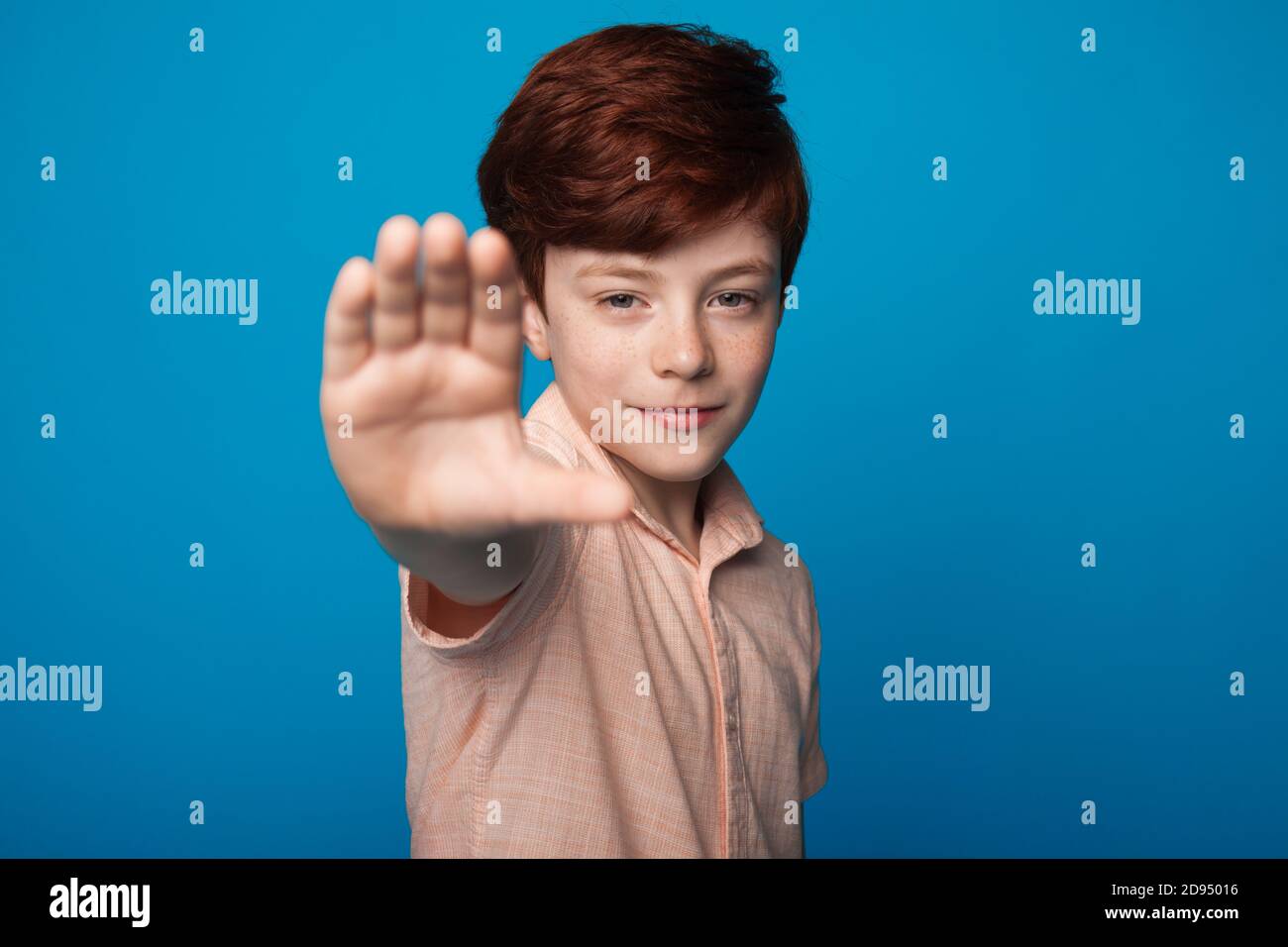 Close up photo of a smiling boy with red hair and freckles gesturing stop sign on a blue wall looking at camera Stock Photo