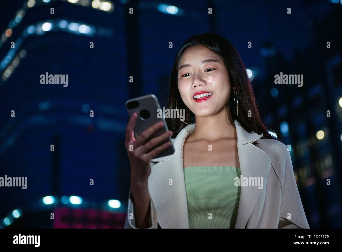 Young pretty Asian woman using mobile phone in the city at night with building bokeh light in background Stock Photo