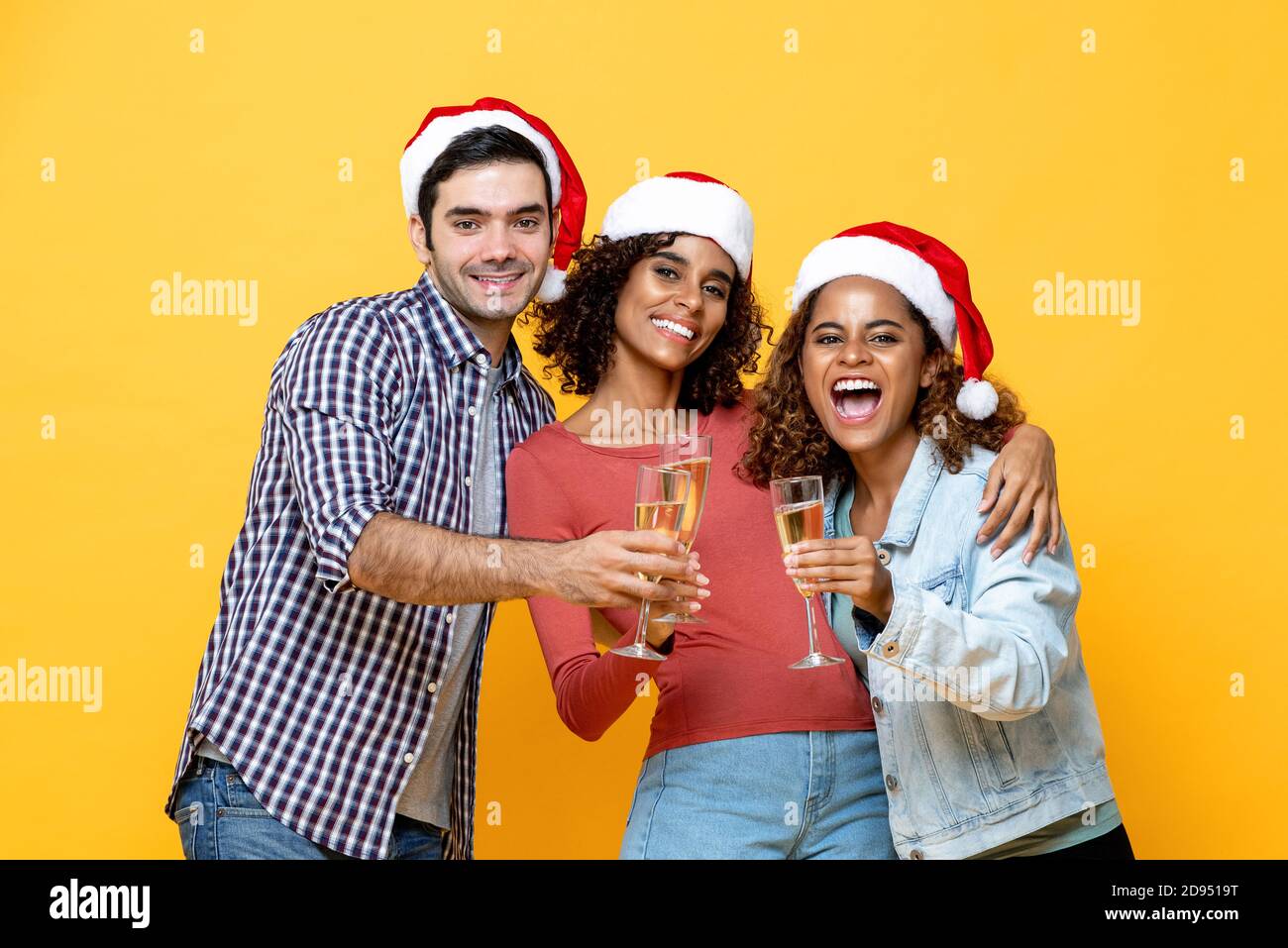 Group of three excited diverse friends celebrating Christmas drinking champagne together on yellow studio background Stock Photo
