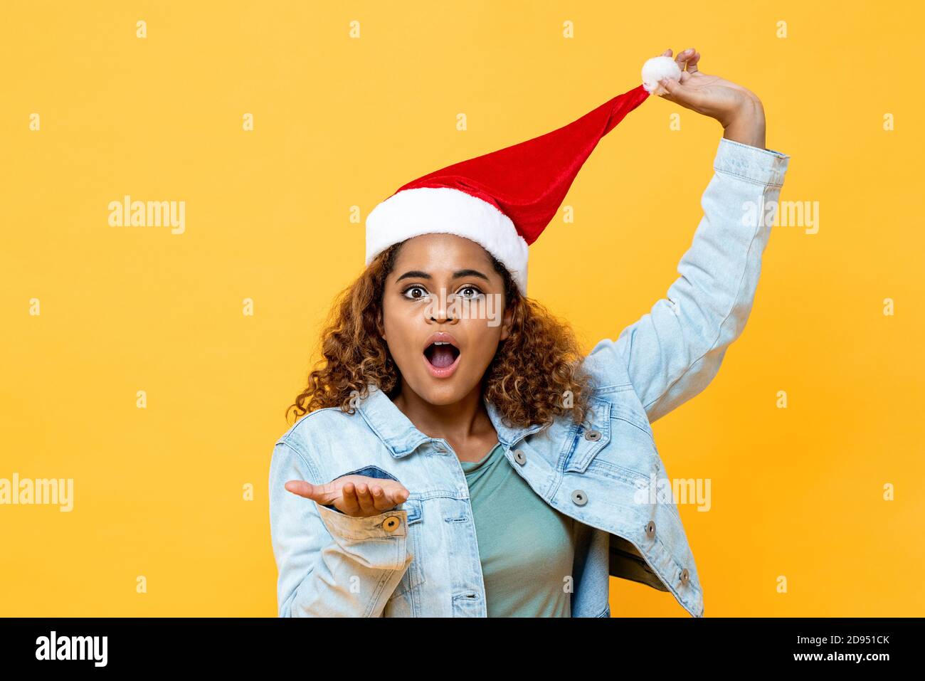 Funny surprised African American woman wearing Christmas hat with open palm gesture on yellow isolated background Stock Photo