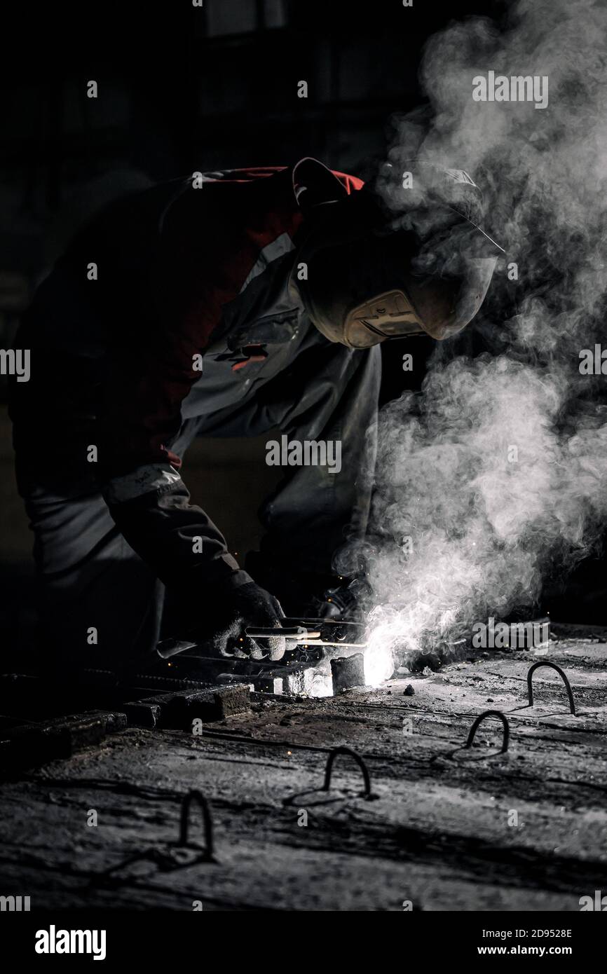 A young  man welder in  uniform, welding mask and welders leathers, weld  metal  with a  welding machine at the construction site, white smoke Stock Photo