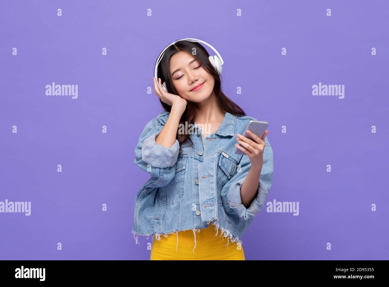 Happy Asian girl wearing headphones listening to music from mobile phone relaxing with eyes closed on isolated purple studio background Stock Photo