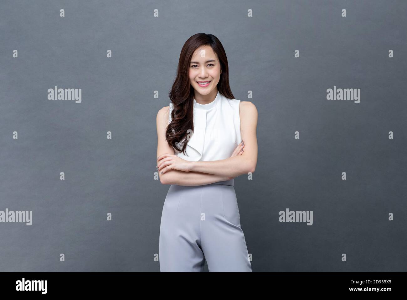 Portrait of smiling young beautiful Asian businesswoman looking at camera and doing arm crossed gesture in isolated studio gray background Stock Photo