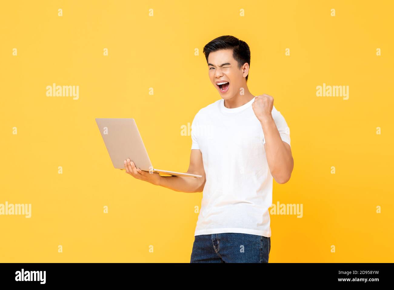 Portrait of excited handsome young Asian man carrying laptop computer and raising his fist doing yes gesture isolated on colorful yellow studio backgr Stock Photo