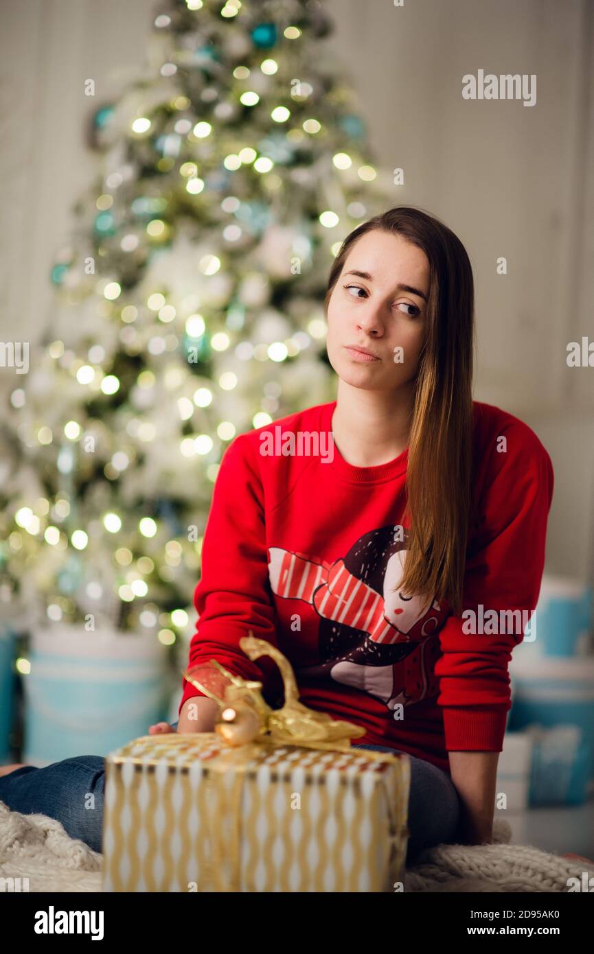 Portrait of beautiful woman with gift sitting by Christmas tree at home Stock Photo