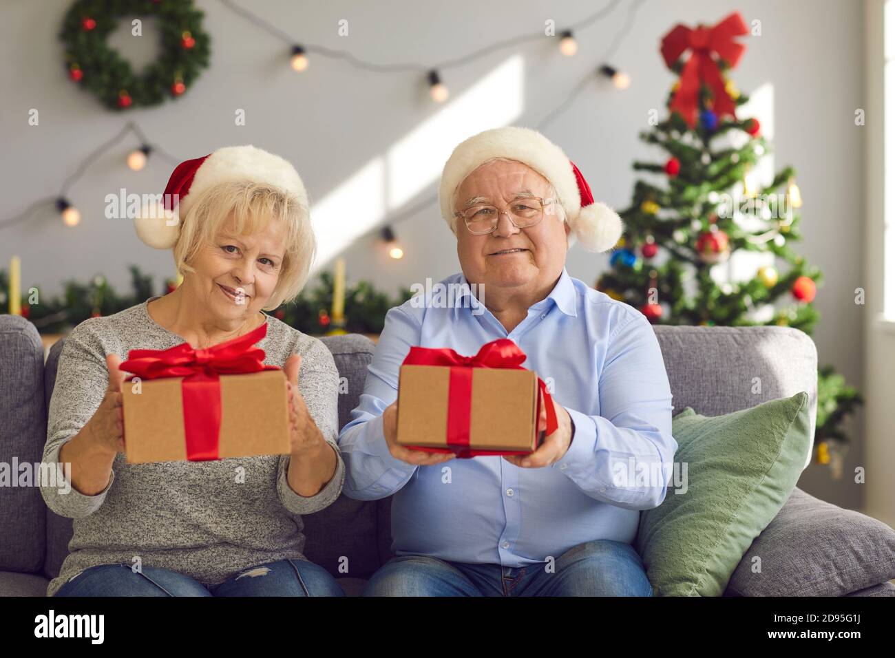Happy grandma and grandma in Santa hats sitting on sofa holding Christmas presents for grandkids Stock Photo