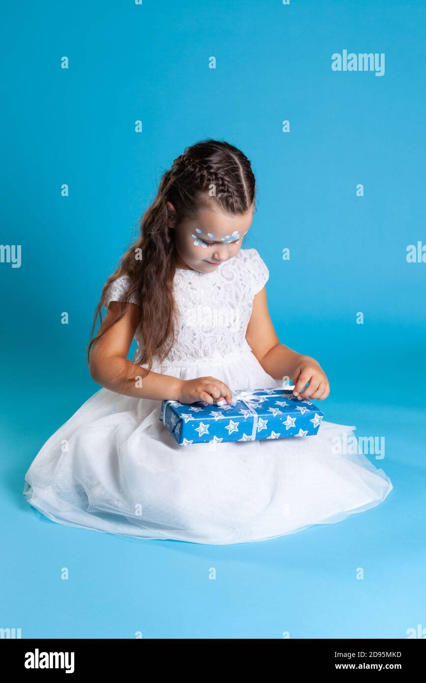a little Princess in a white dress sits on her lap and unwraps a Christmas gift, isolated on a blue background Stock Photo