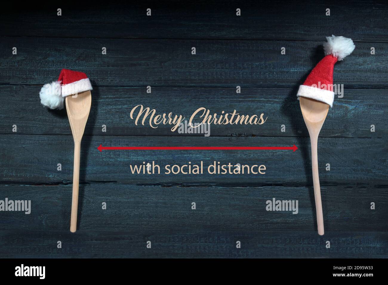 Two cooking spoons with red Santa Claus hats, between them text Merry Christmas with social distance, dark blue wooden background, health safety conce Stock Photo