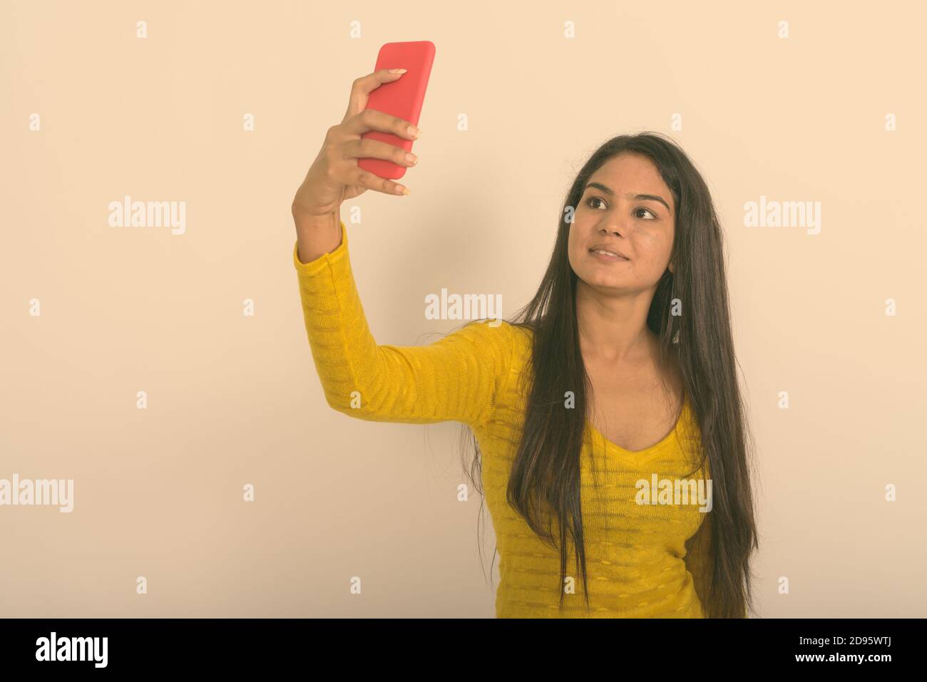 Studio shot of young happy Indian woman smiling while taking selfie picture with mobile phone against white background Stock Photo