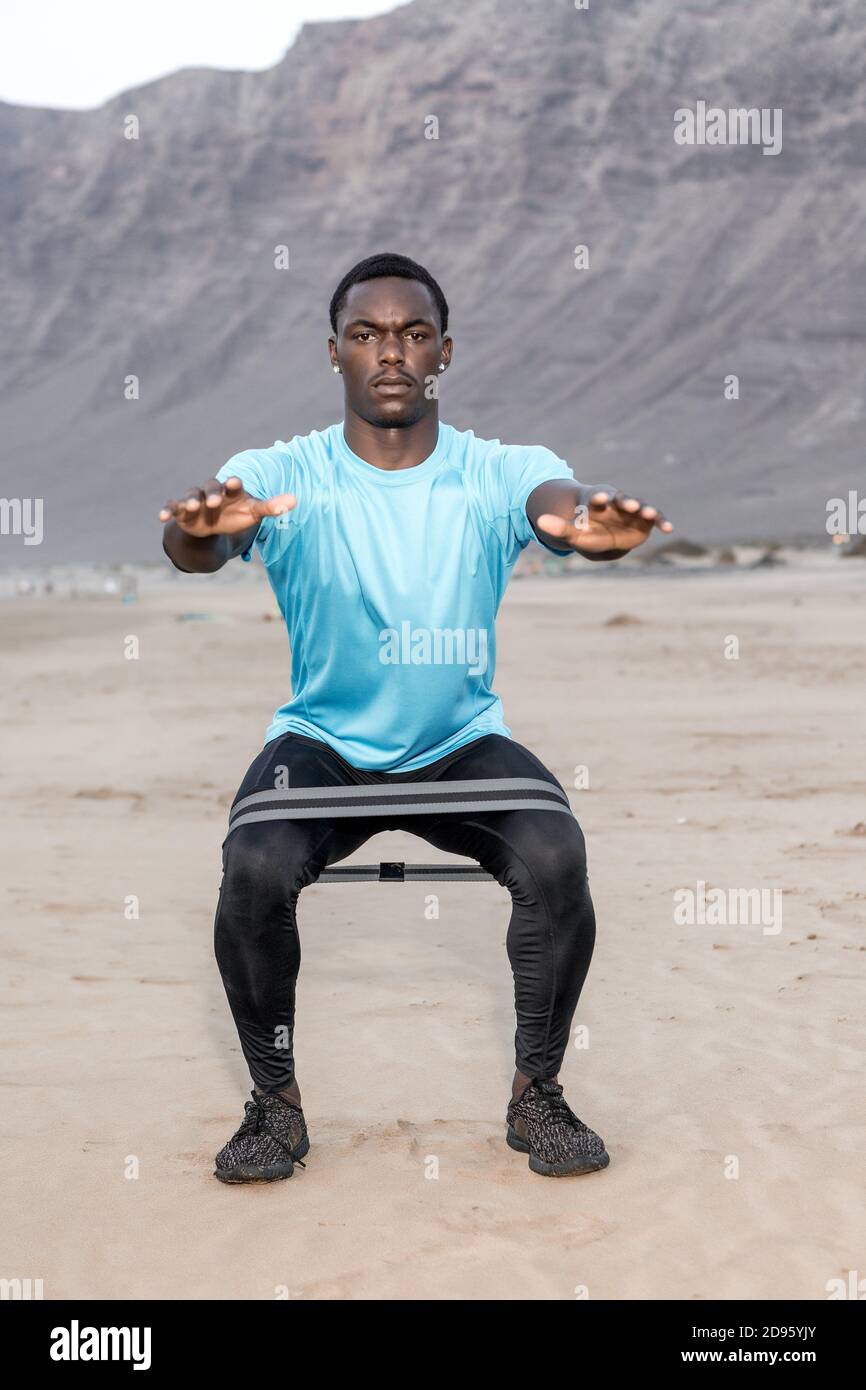 Full body serious African American sportsman squatting with rubber band during fitness workout on Famara Beach in Lanzarote, Spain. Stock Photo