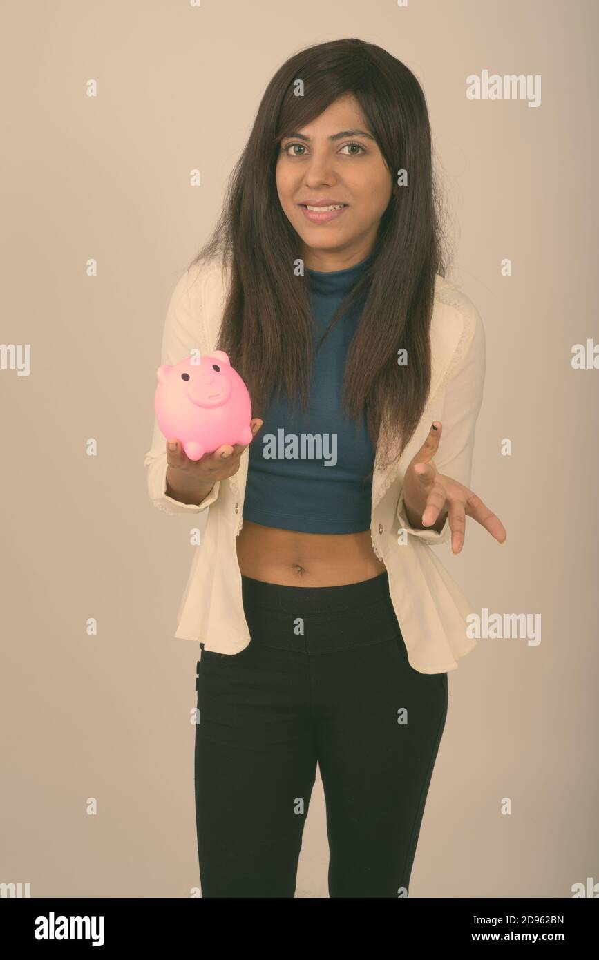 Young happy Persian businesswoman smiling while looking confused and holding piggy bank Stock Photo
