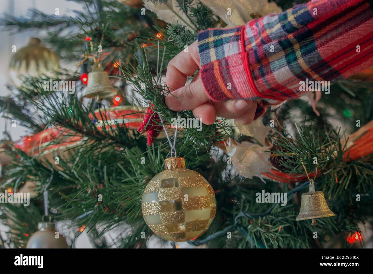 Girl in red pajamas placing a decorative ball on the Christmas tree. Closeup girl holding Christmas ball preparing the Christmas tree for this 2020. Stock Photo