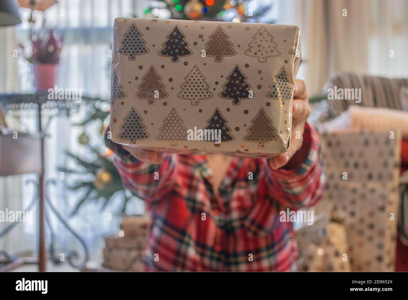 Young girl with Christmas pyjama holds a present for her boyfriend in a Christmas Day. Winter Photography 2020. Christmas Tree Concept. Stock Photo
