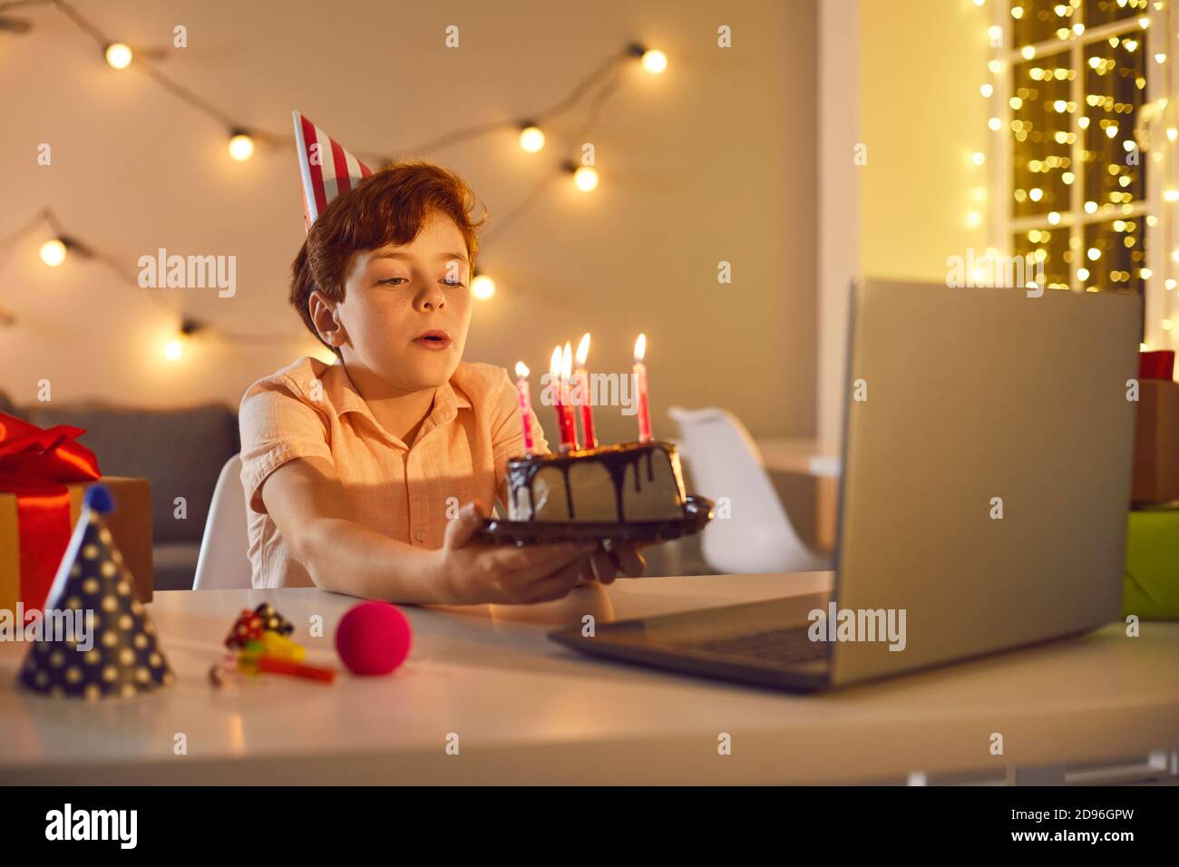 Little birthday boy making a wish and blowing burning candles on cake during online party at home Stock Photo