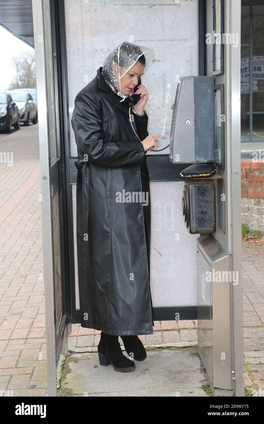 Older woman in long black coat, looking worried & concerned with rain mate on public telephone. Calling the emergency service, calling  friend in dirty telephone box, kiosk. Stock Photo