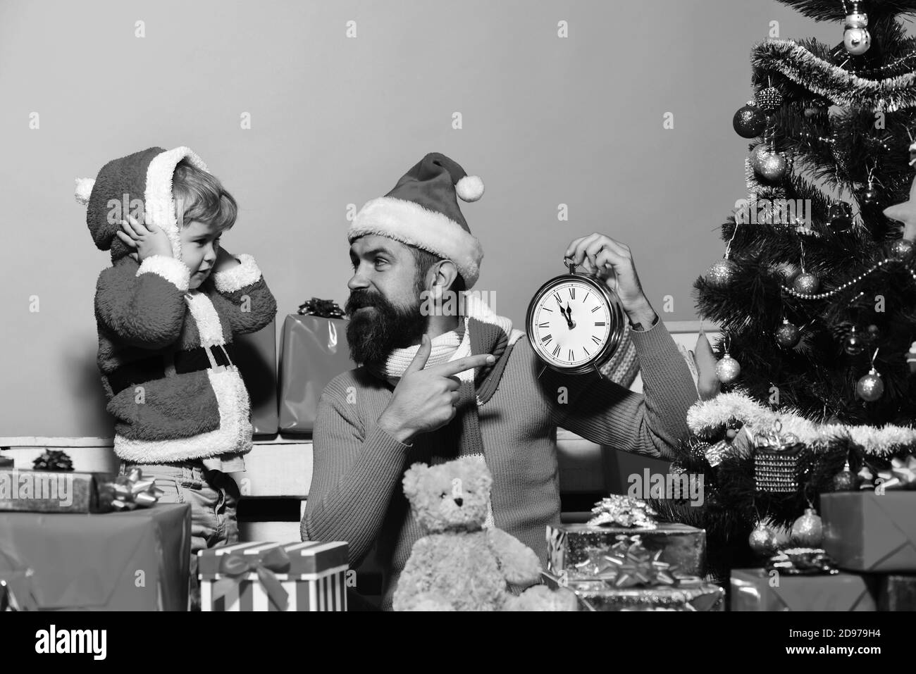 Holiday and countdown concept. Santa and little assistant close ears and point at ringing alarm clock. Christmas family waits for New Year on blue background. Boy and man with beard and serious faces Stock Photo