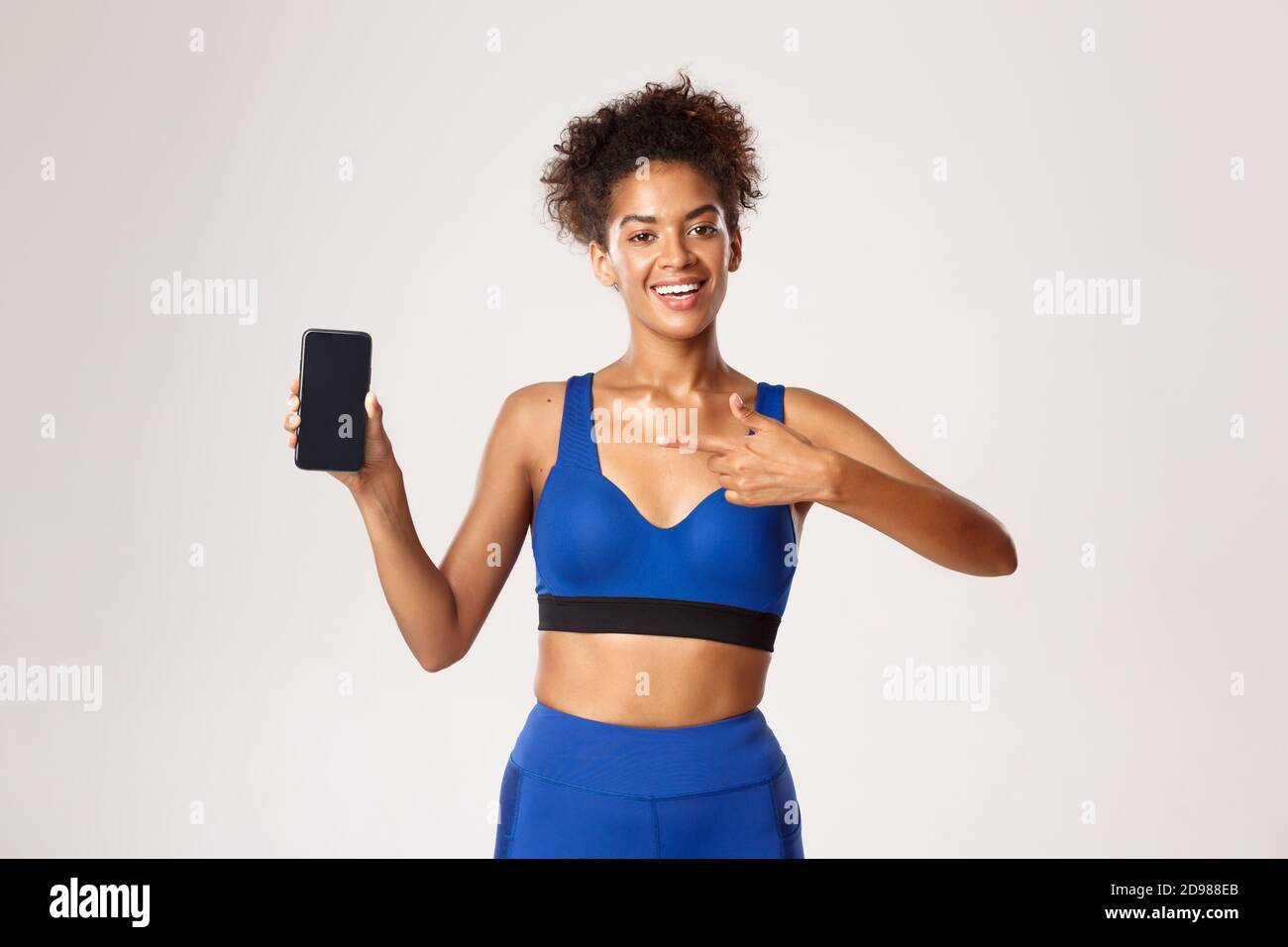 Sport and technology concept. Beautiful athletic woman in fitness clothing, pointing finger at smartphone screen, standing against white background Stock Photo