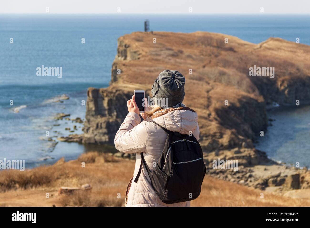 A young woman admires a beautiful view of the sea and rocks at Cape Tobizin on the Russky Island in Vladivostok on a sunny autumn day. Stock Photo