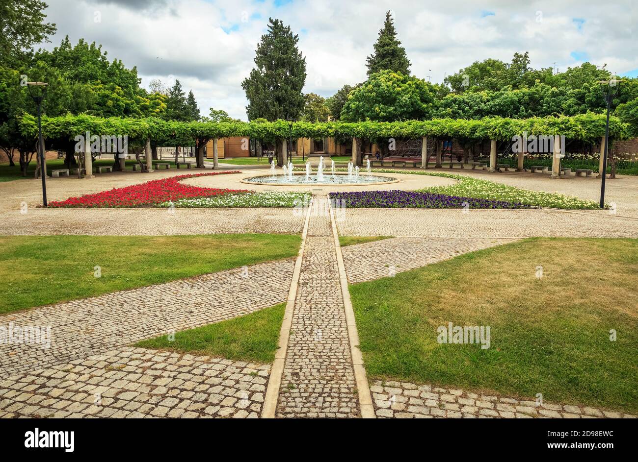 Landscape of the Castelo Branco city park in Portugal, with the lake and fountain framed by an arbor and flower beds, on a spring day with clouds. Stock Photo