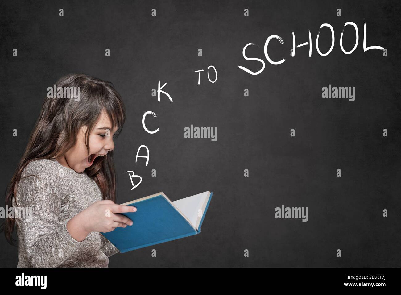 Cute tanned long haired  little girl is surprised at the letters Back to School coming out of an open book she is holding in her hands. Stock Photo