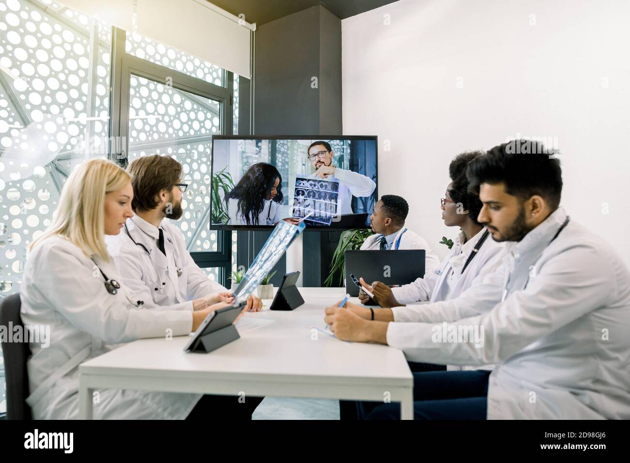 Front view of five multiracial confident doctors discussing treatment plan of patient while having video conference meeting with their Afrcian and Stock Photo