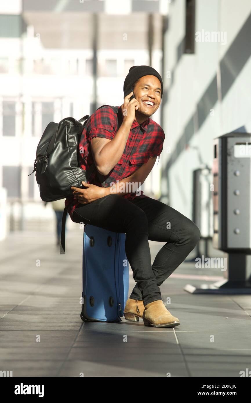 Portrait of happy travel man sitting on suitcase at station while talking on cellphone Stock Photo