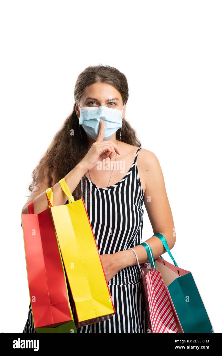 Adult woman making shush secret gesture with index finger over mouth covered by medical or surgical mask holding colourful shopping gift bags isolated Stock Photo