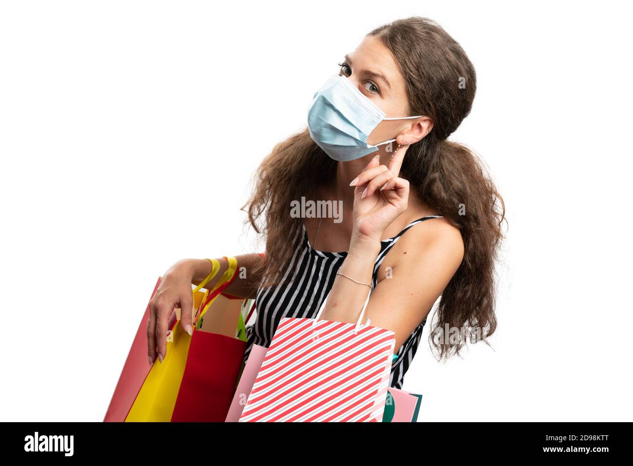 Adult woman model making listening gesture touching ear holding colourful bags wearing mask face covering as covid19 pandemic shopping or consumerism Stock Photo