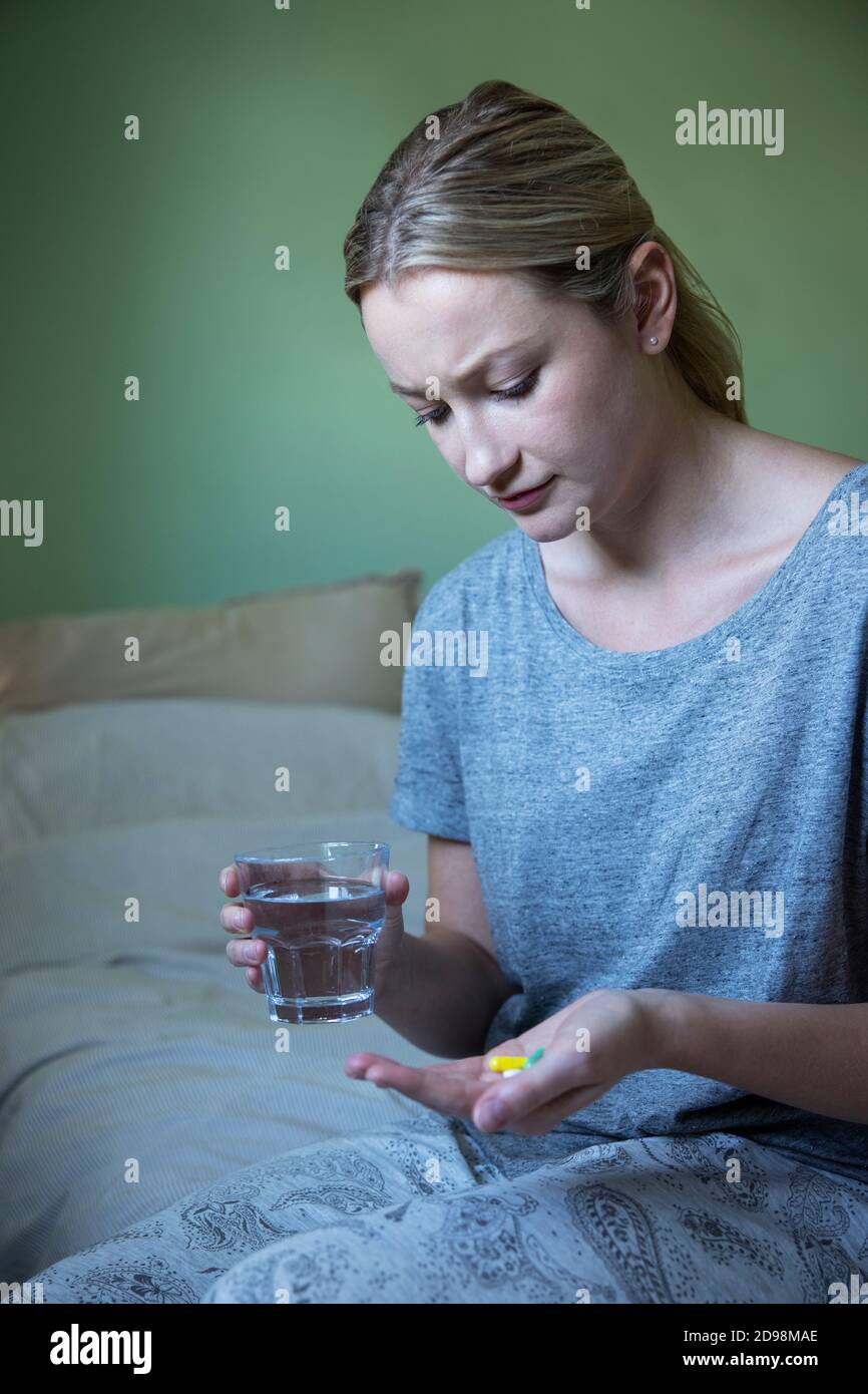Unhappy Woman Sitting On Bed Wearing Pyjamas Taking Medication With Glass Of Water Stock Photo