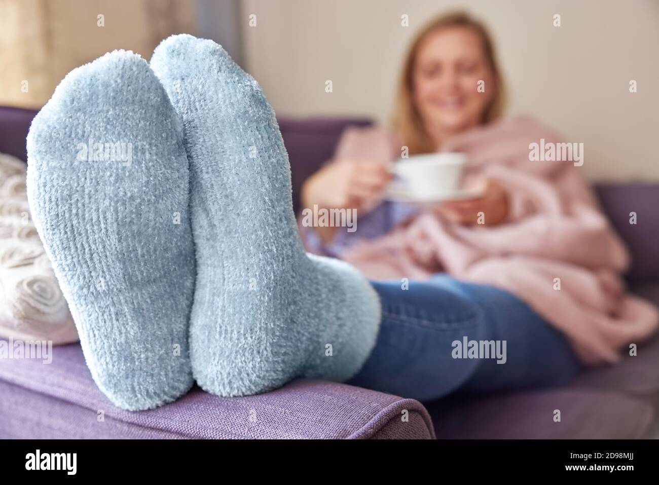 Woman At Home Wearing Cosy Warm Socks And Wrapped In Blanket Lying On Sofa Drinking Cup Of Tea Stock Photo