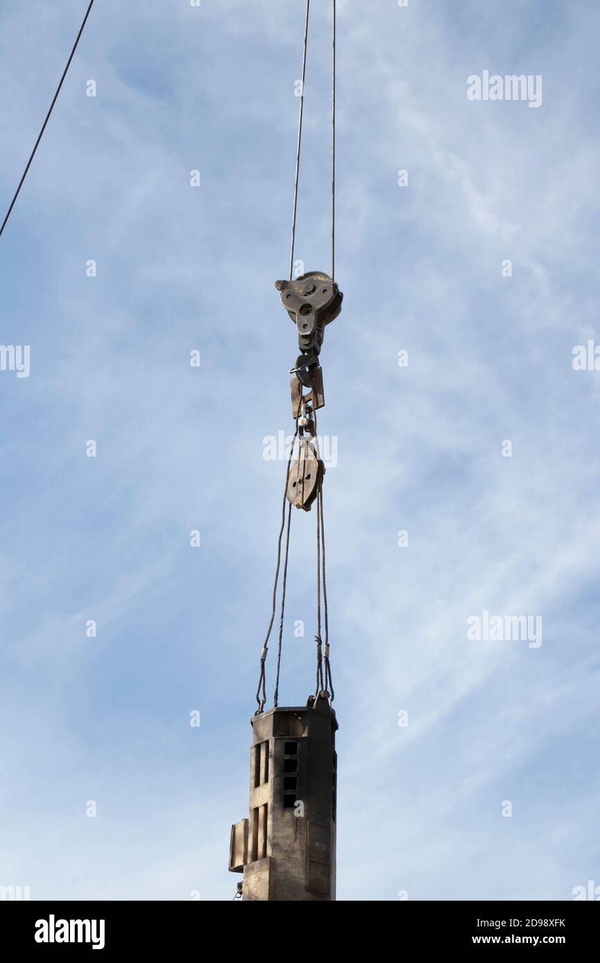 Construction crane with a hook and a large steel industrial spike hanging on ropes lifting up, low angle view with cloudy sky Stock Photo