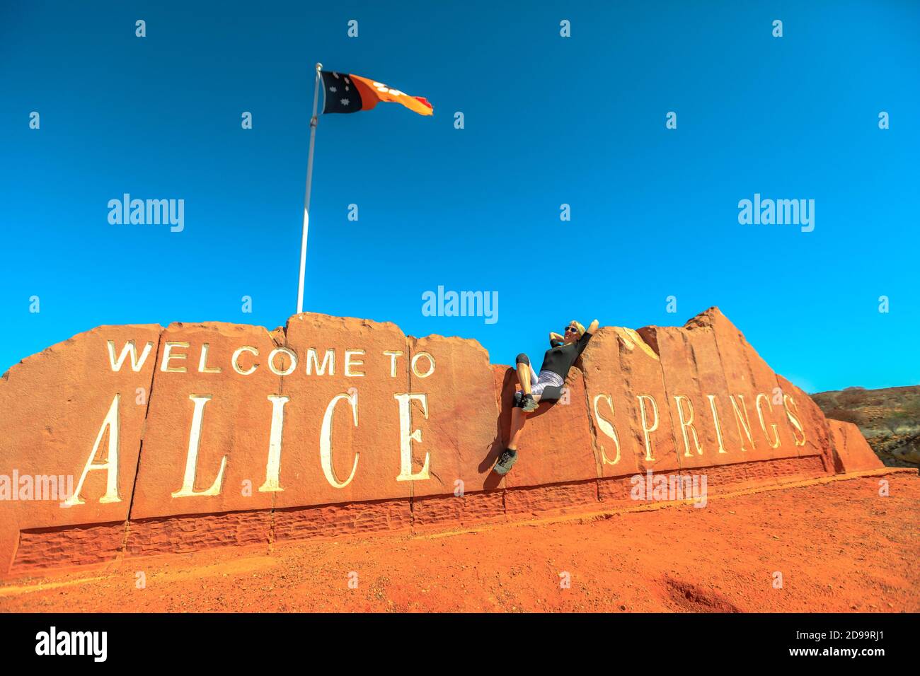 Happy carefree tourist woman at Alice Springs Welcome Sign in Northern Territory, Central Australia. Tourism in Outback Red Center desert. Travel Stock Photo