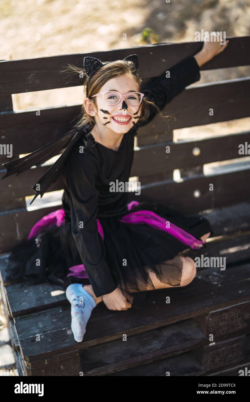 Cute little girl wearing a Halloween cat costume and sitting on a bench ...