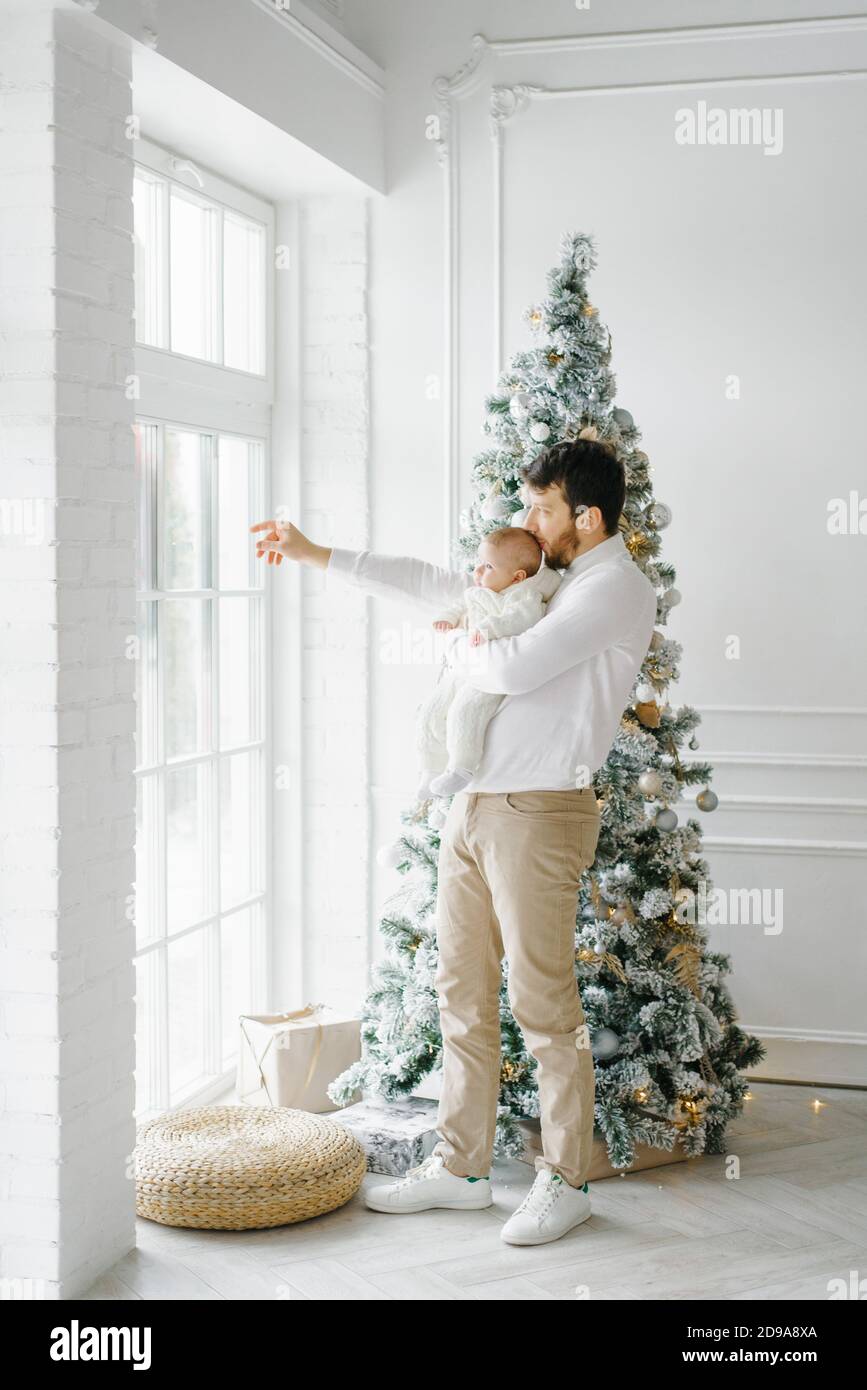 The father holds the baby in his arms and shows him something through the window, standing by the Christmas tree in the white living room. new year ce Stock Photo