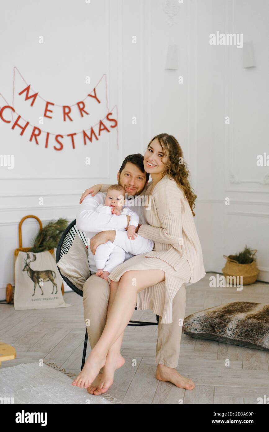 Young parents lift their baby in their arms and smile at him against the background of a bedroom decorated for Christmas and New Year Stock Photo