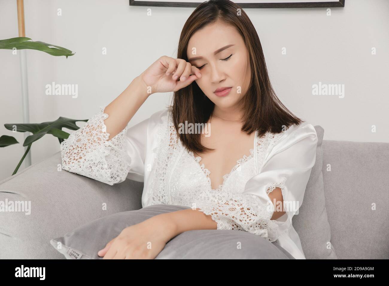 Asian women in a nightgown and wear white satin robe rubbing her eye sitting on the gray sofa in the living room at night, A girl in silk nightwear sc Stock Photo