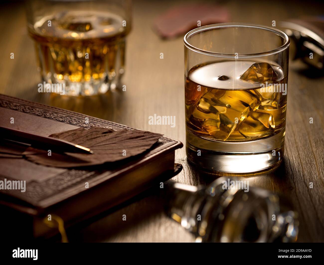 Backlit glass of whisky on the rocks on a wooden table, with notebook, pen and a second glass Stock Photo