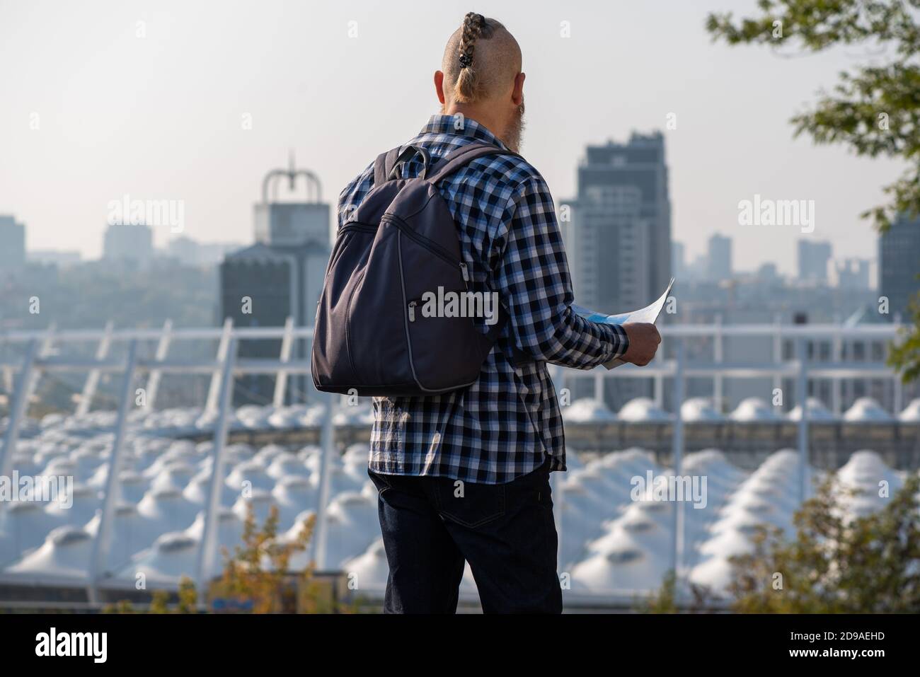 Bearded caucasian man tourist in plaid shirt with backpack on his back looking at map planning route in the city center. Rear view Stock Photo