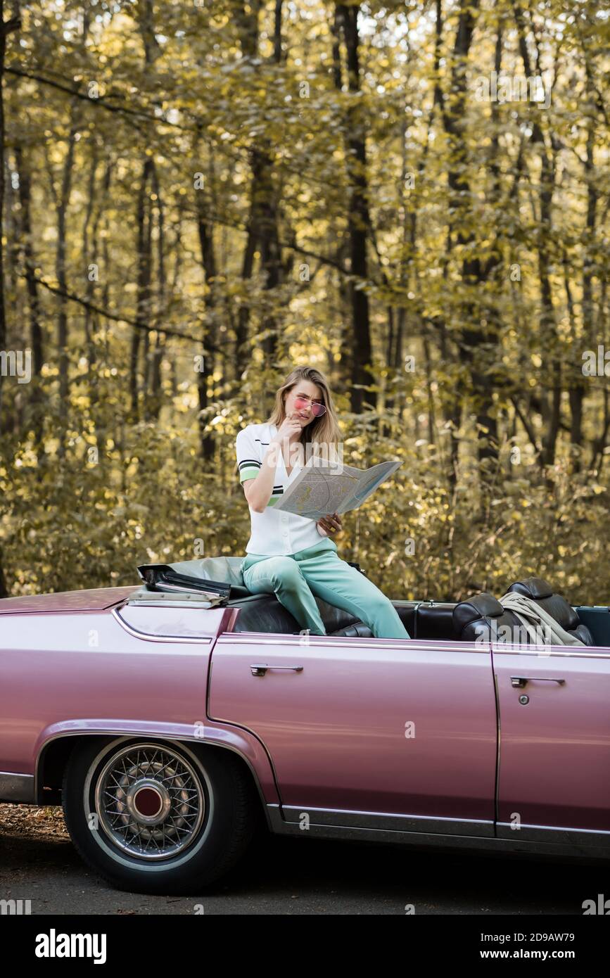 thoughtful woman in sunglasses looking at map while sitting in cabriolet in forest Stock Photo