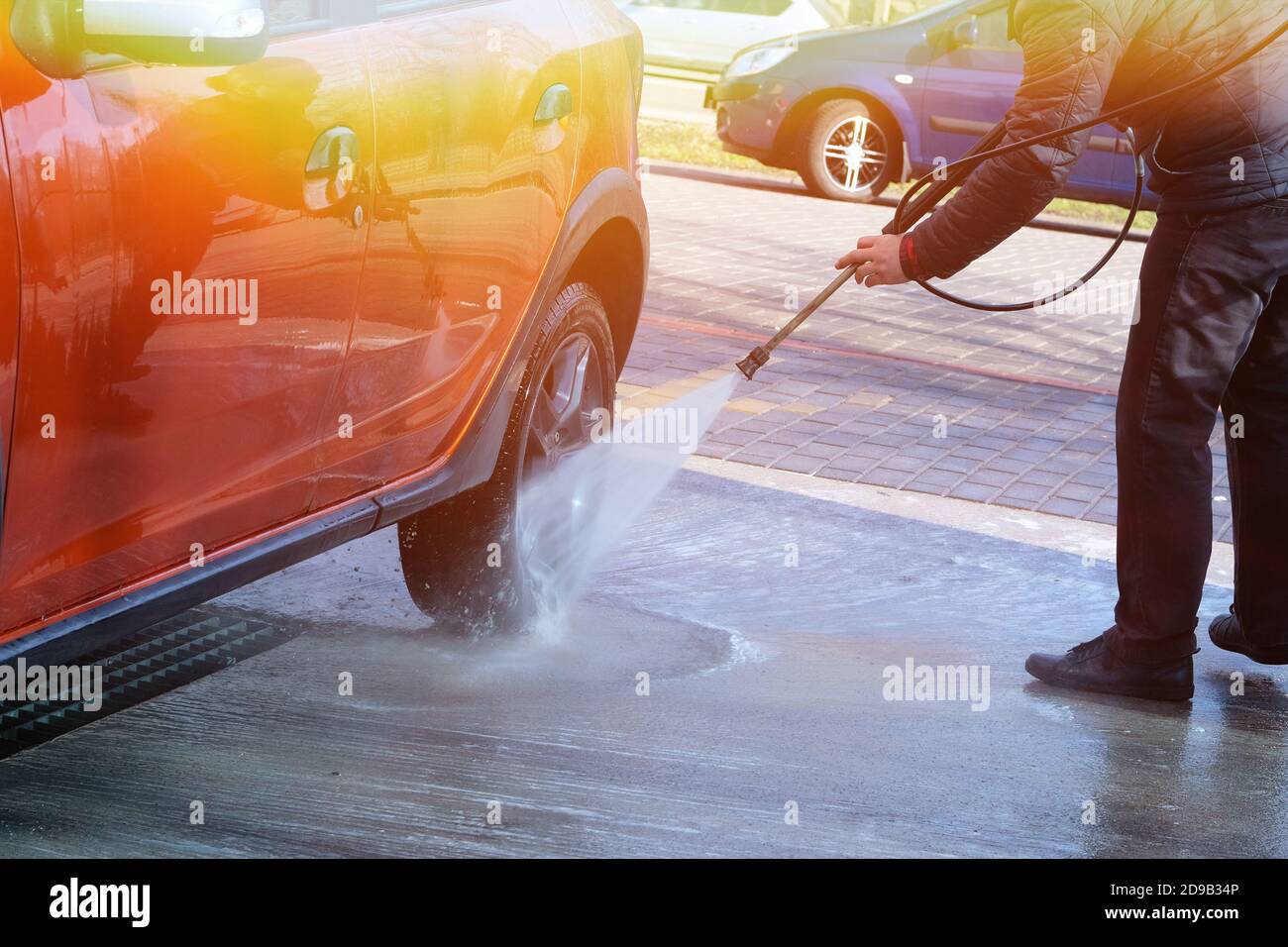 Man washes his orange car at car wash in outdoors. Cleaning with water at self-service car wash. Sunlight. Stock Photo