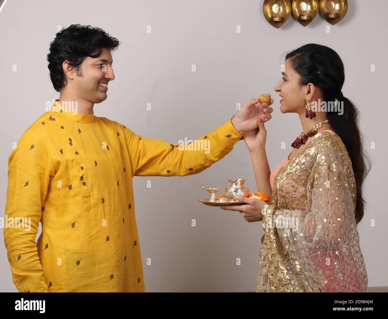 Indian smart couple eating sweet laddu on karva chauth Diwali or anniversary, selective focus. while girl or wife holding puja thali. Stock Photo