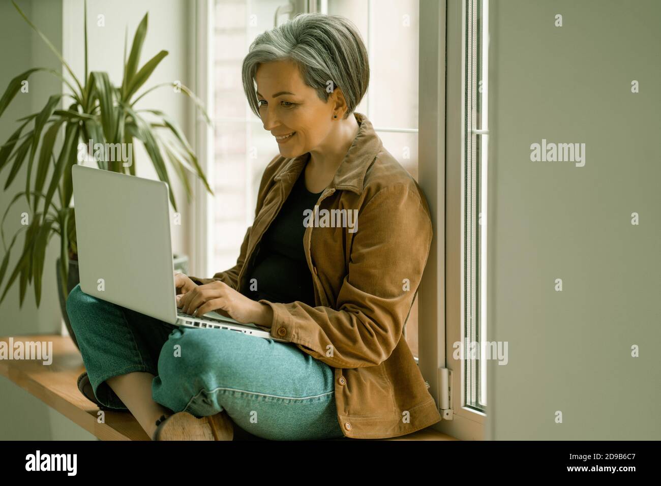Smiling mature woman looks at web camera taking part in virtual video chat or working with computer or while sitting with her back against window Stock Photo
