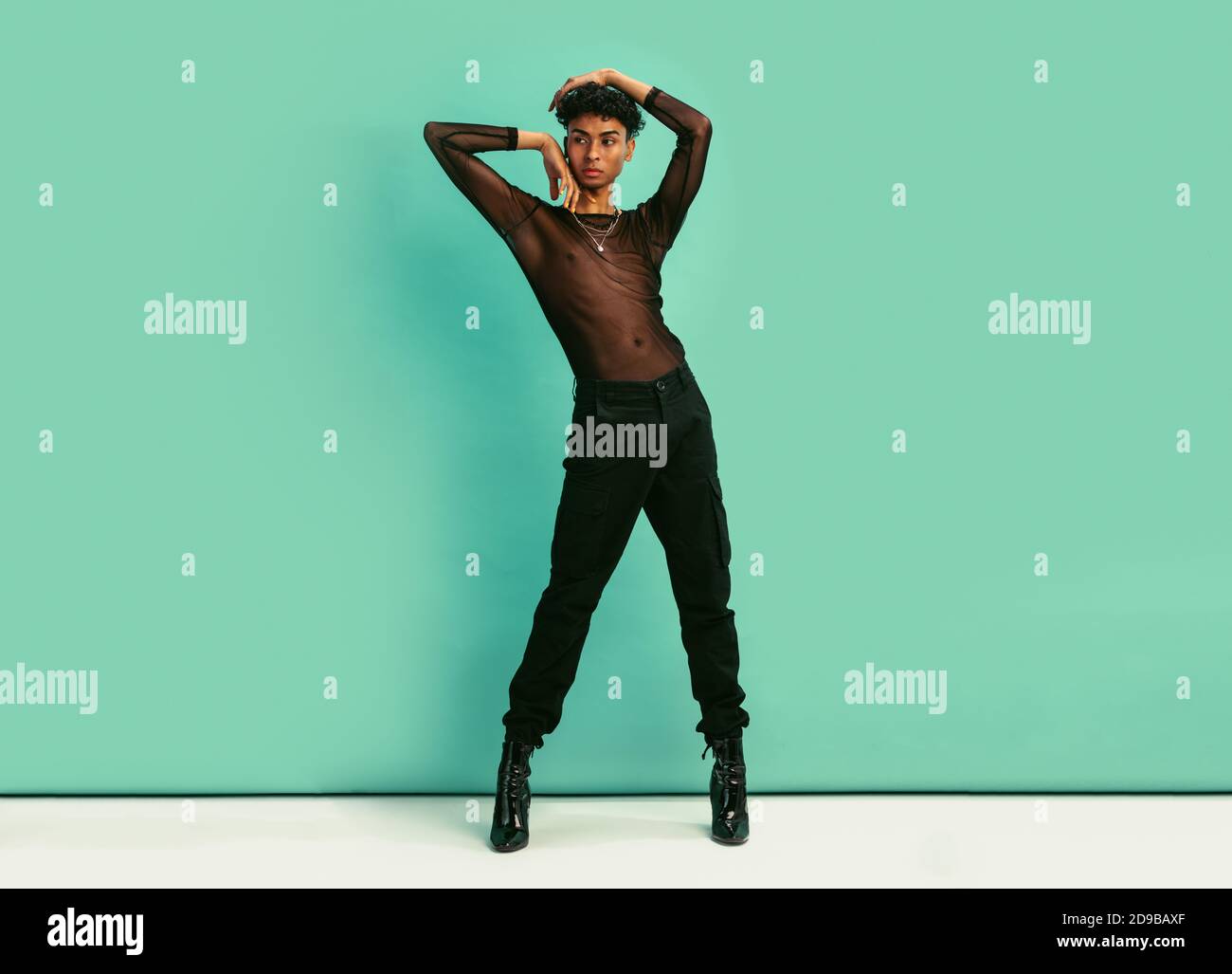 Full length of a gay man in black outfit. Gender fluid man wearing black jeans, black net shirt and high heels posing against blue background. Stock Photo