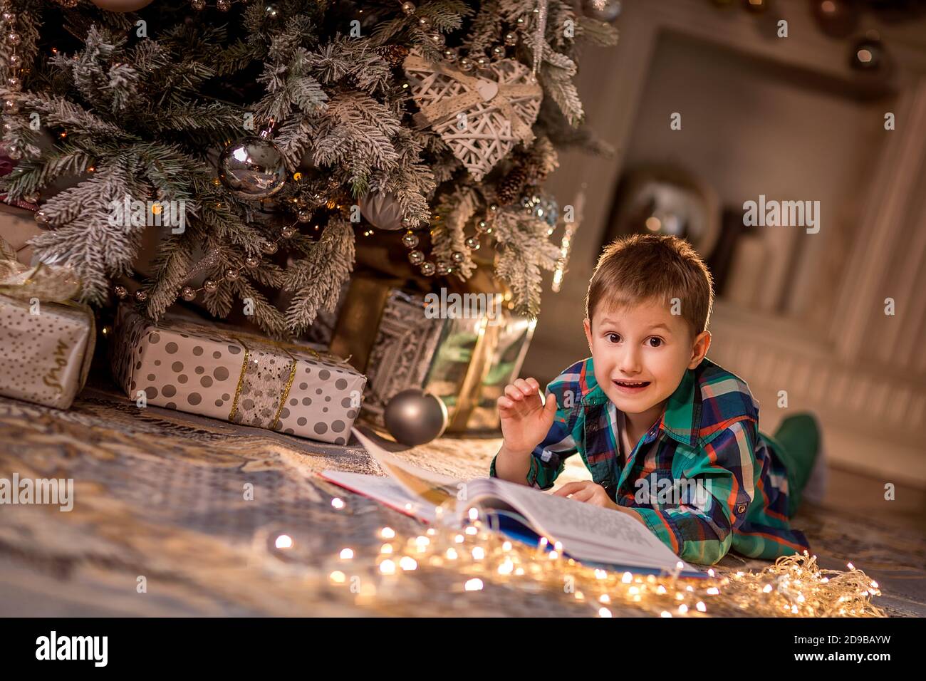 The boy is lying on the floor near a decorated Christmas tree. Reading a book with Christmas stories. Cozy winter evenings at home. Concept of Christm Stock Photo