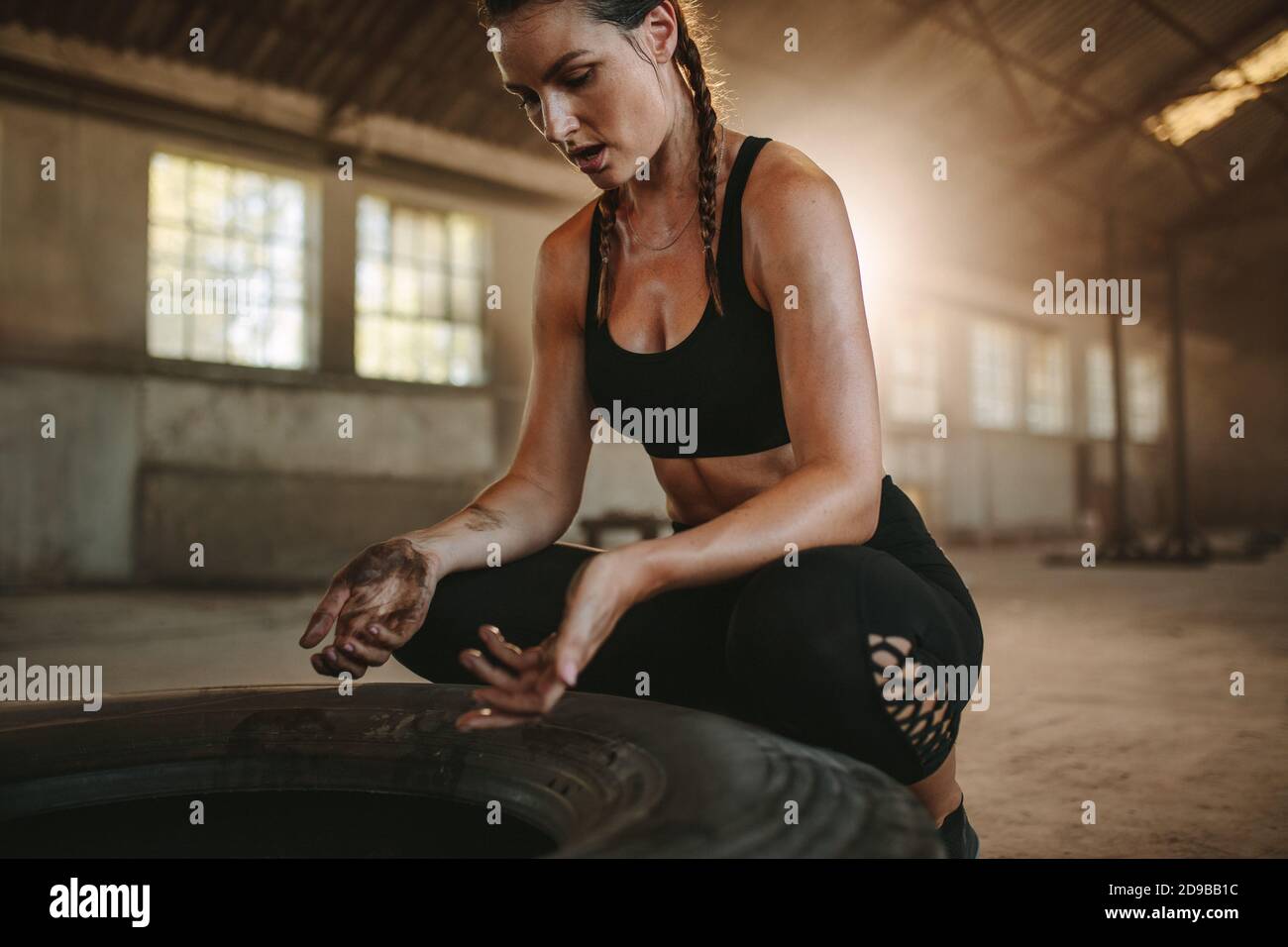 Muscular female athlete taking a break from cross training workout. Woman in black sportswear sitting by big tire in empty warehouse. Stock Photo